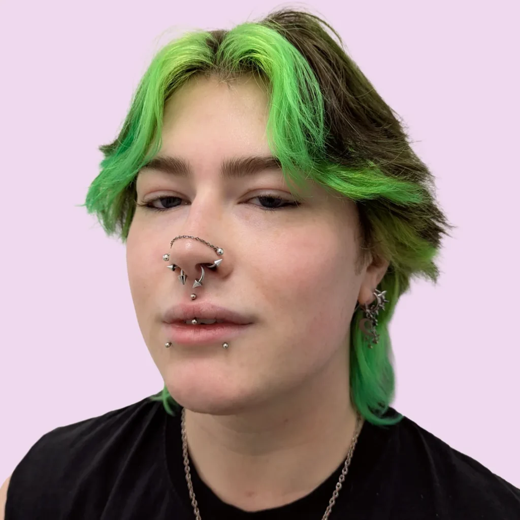 Portrait of a person with green hair and multiple facial piercings, including a septum, snakebites and ear piercings